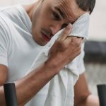Active Recovery - Young male wiping forehead with towel during rest after hard workout in contemporary fitness center
