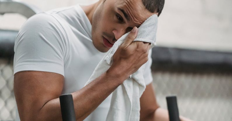Active Recovery - Young male wiping forehead with towel during rest after hard workout in contemporary fitness center
