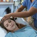 Recovery Tools - Crop African American female doctor with professional equipment doing examination of ear of woman lying on bed in hospital ward